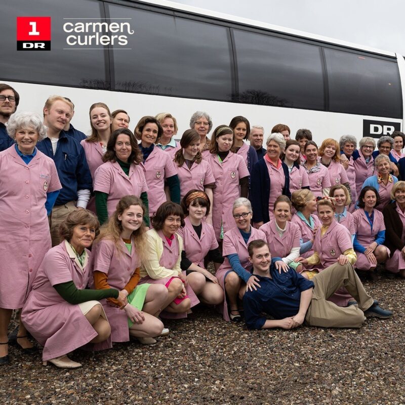 Group photo of the cast in pink factory uniforms in front of the DR bus on the set of Carmen Curlers