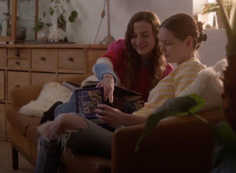 Still from the film Heltinde where Sille Schnoor sits on a sofa reading a book with a girl