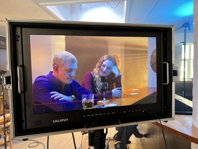 Camera monitor shows two actors at a table in a scene during filming