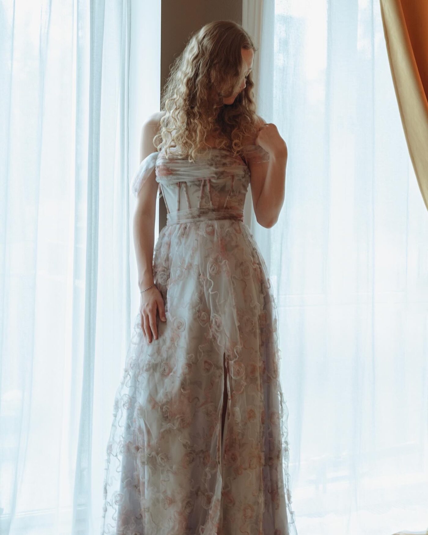 Sille Schnoor in a long floral dress by a light curtain with her back half to the camera