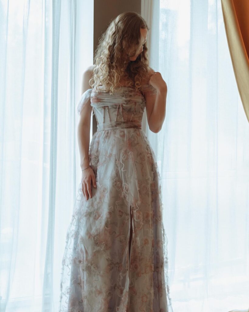 Sille Schnoor in a long floral dress by a light curtain with her back half to the camera