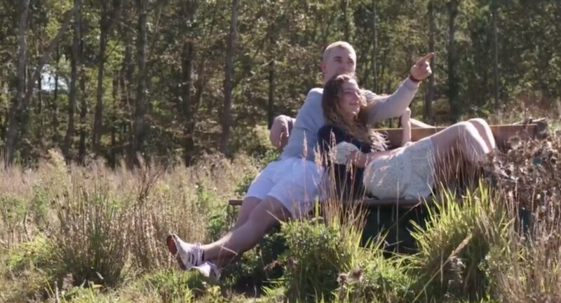 Film still from an outdoor scene where a man points out across a heather-covered landscape and a woman in a white dress lies on a bench