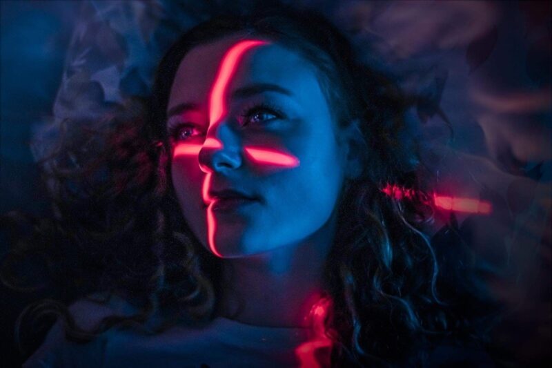 Sille Schnoor lies with her eyes closed while red neon stripes appear across her face in blue light.