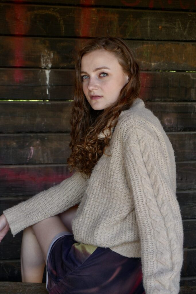 Sille Schnoor sits against a dark wooden wall in a light knitted blouse and looks into the camera