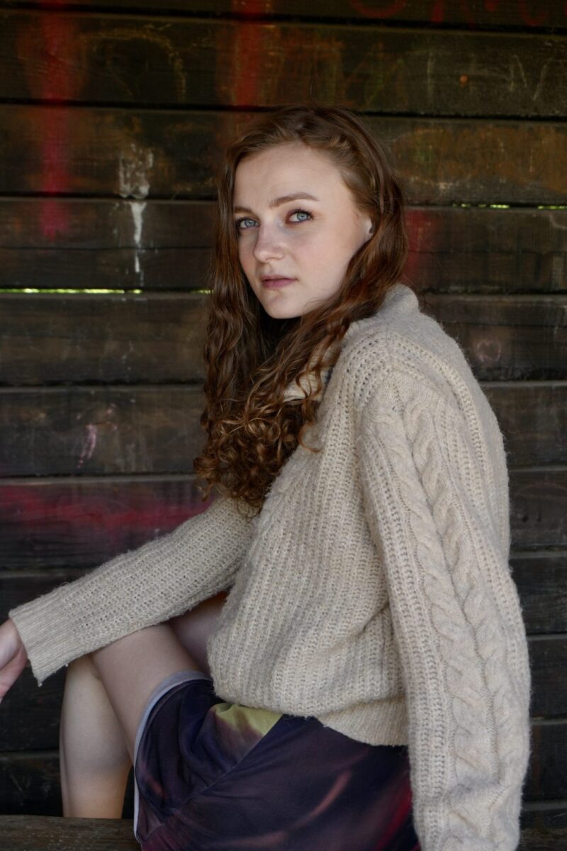 Sille Schnoor sits against a dark wooden wall in a light knitted blouse and looks into the camera