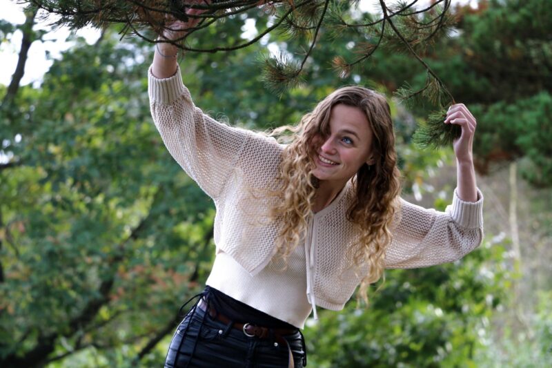Sille Schnoor smiles and reaches up for a pine branch in a white knitted cardigan