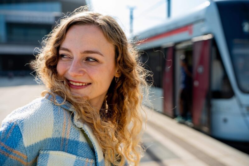 Sille Schnoor smiles over her shoulder at a light rail station in a blue checkered jacket
